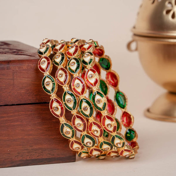A gold-plated bangle bracelet decorated with red, green, and white enamel motifs, draped over a dark wooden box with a brass incense burner in the background.
